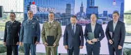 Sześciu mężczyzn stoi na tle ekranu z logotypem PKO Banku Polskiego i panoramą Warszawy. Trzech z nich ma na sobie mundury wojskowe i policyjne, pozostali garnitury. Każdy trzyma w ręku teczkę lub dokument. To moment wspólnej prezentacji po podpisaniu umowy lub porozumienia.