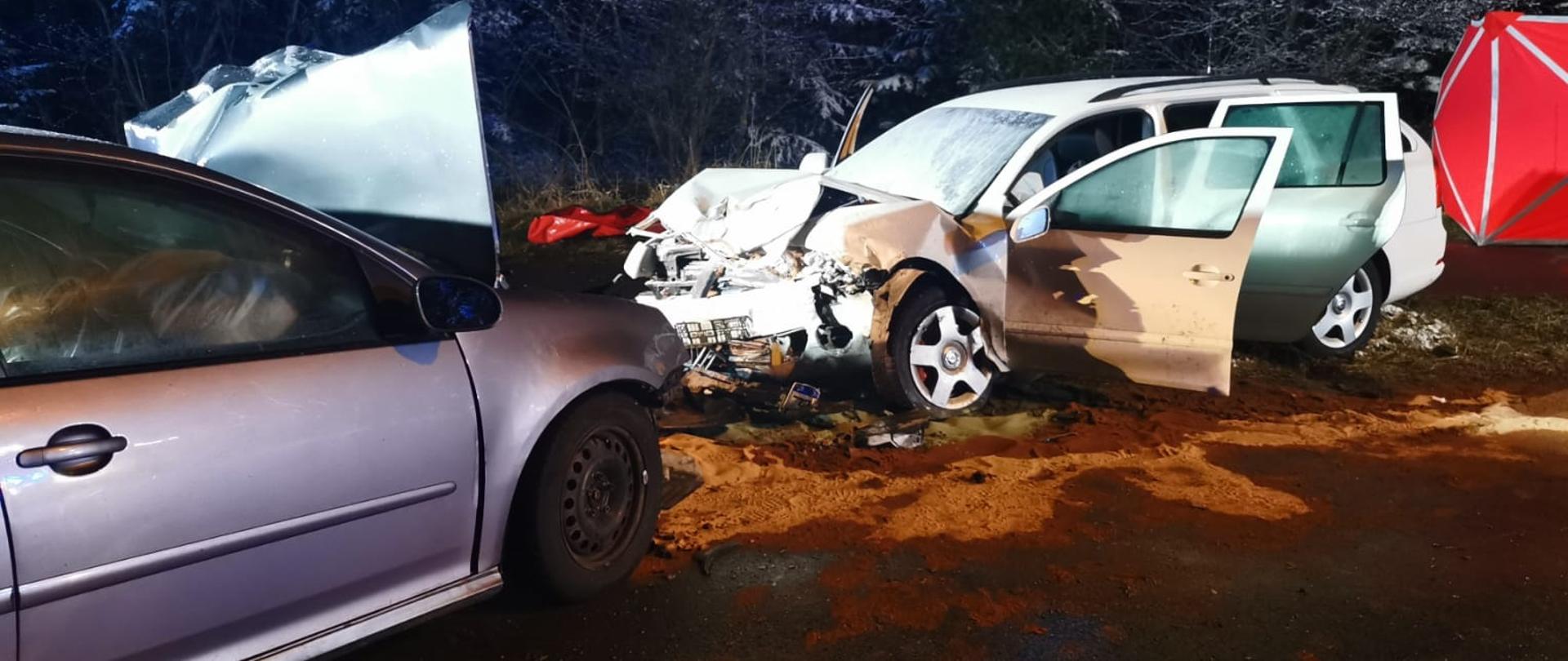 Dwa samochody bardzo mocno uszkodzone stoją na jezdni.