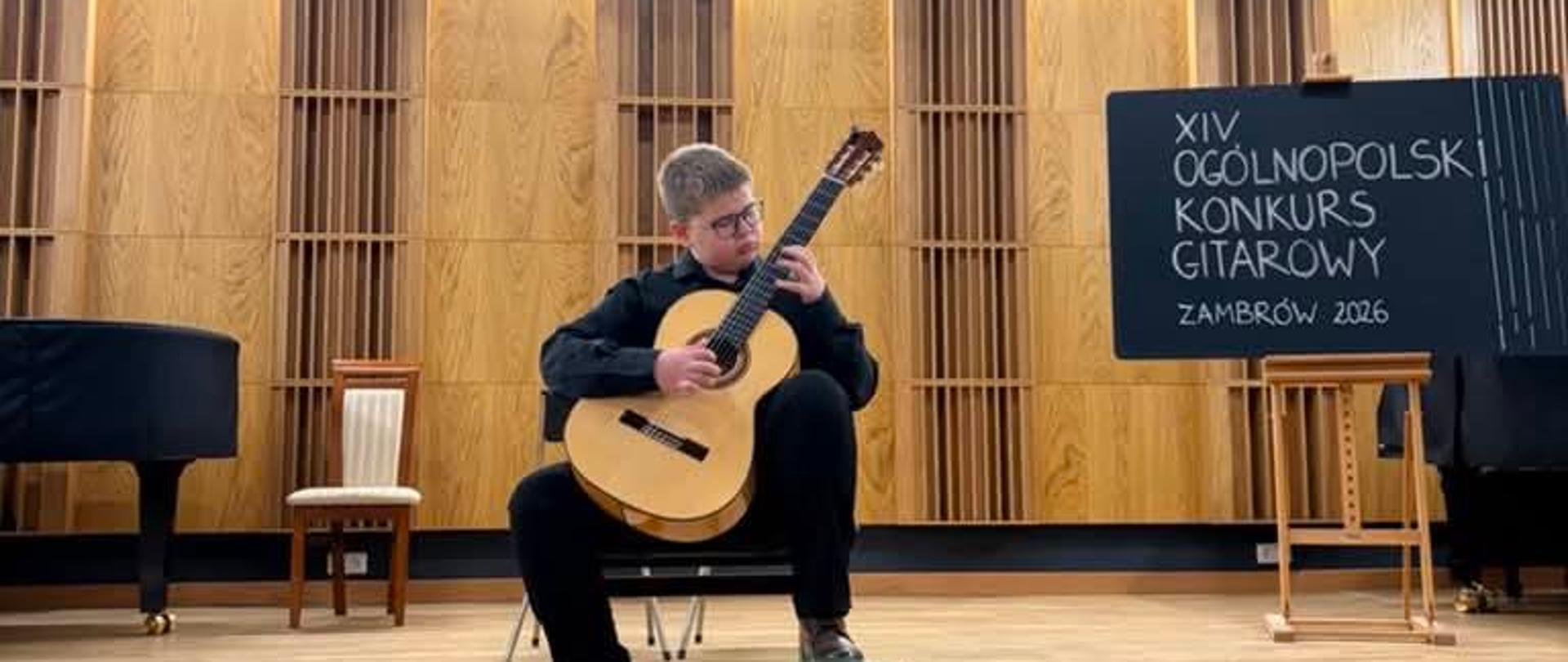 Na scenie siedzi chłopiec i gra na gitarze klasycznej. Chłopiec ubrany jest w czarne spodnie i czarną koszulę. Z jego prawej strony tablica z napisem, czternasty Ogólnopolski Konkurs Gitarowy Zambrów 2026. 