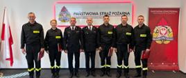 Zdjęcie przedstawia strażaków biorących udział w uroczystości. W tle znajduje się napis Państwowa Straż Pożarna, po lewej stronie znajduje się flaga biało czerwona a po prawej znajduje się rollup z napisem Państwowa Straż Pożarna i logiem PSP