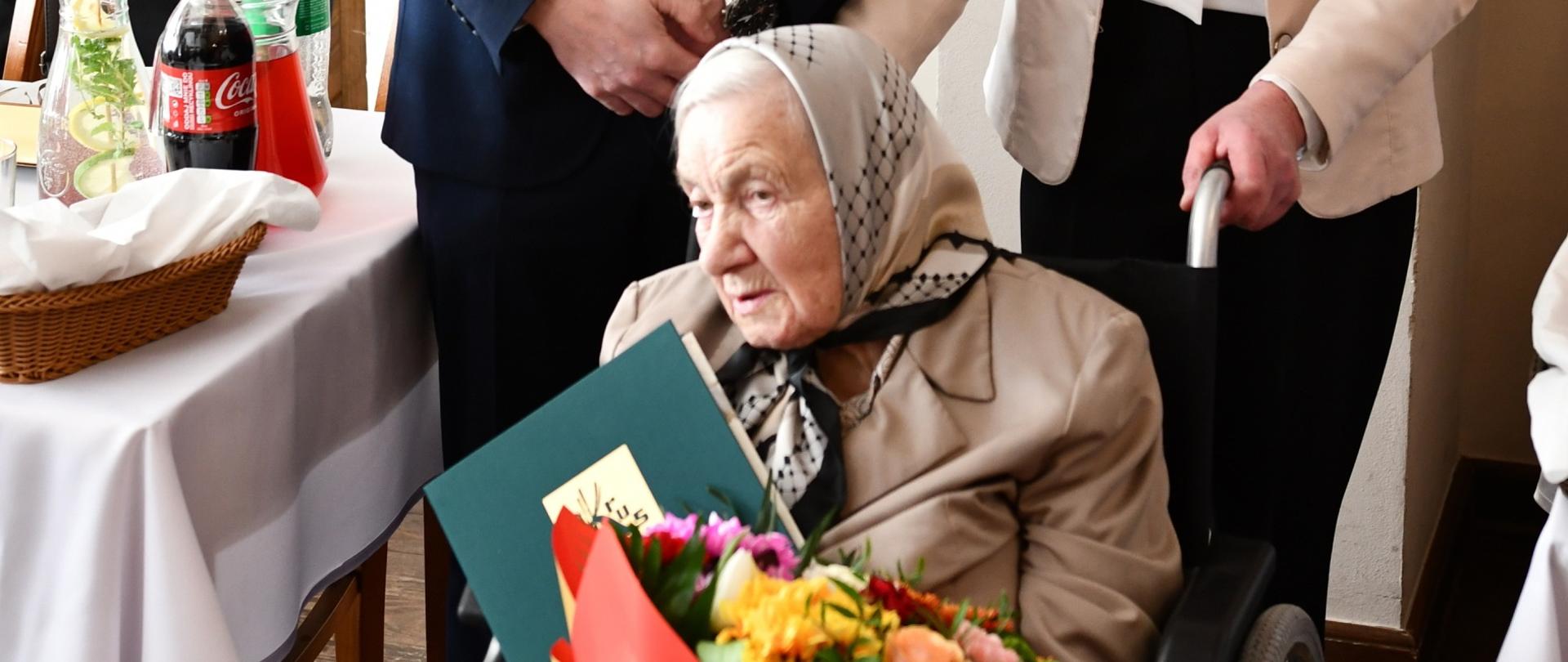 Starsza kobieta siedzi na wózku inwalidzkim z bukietem kwiatów i listem gratulacyjnym w zielonej okładce ze złotym logo KRUS, za nią stoją mężczyzna i kobieta