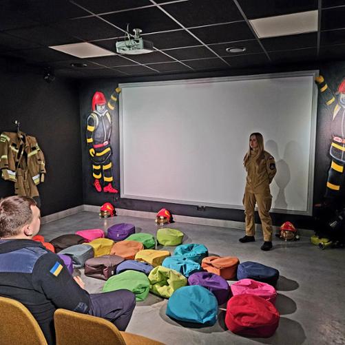 Zdjęcie pokazuje strażaczkę podczas prezentacji. Na podłodze leżą kolorowe poduszki do siedzenia , na ścianach są rysunki strażaków. 