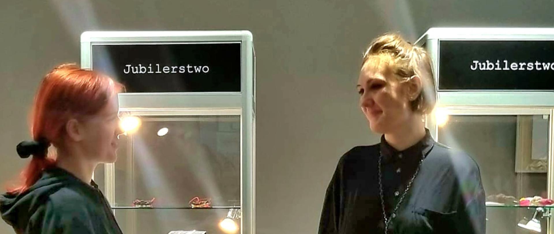 Fotografia przedstawiająca dwie osoby - jedną z nich jest autorka wystawy biżuterii pn. Materia - dr Michalina Owczarek-Siwak, w tle widoczne są witryny wystawiennicze.