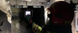 Na pierwszym planie widać strażaka z kamerą termowizyjną sprawdzającego temperaturę ściany spalonego mieszkania.