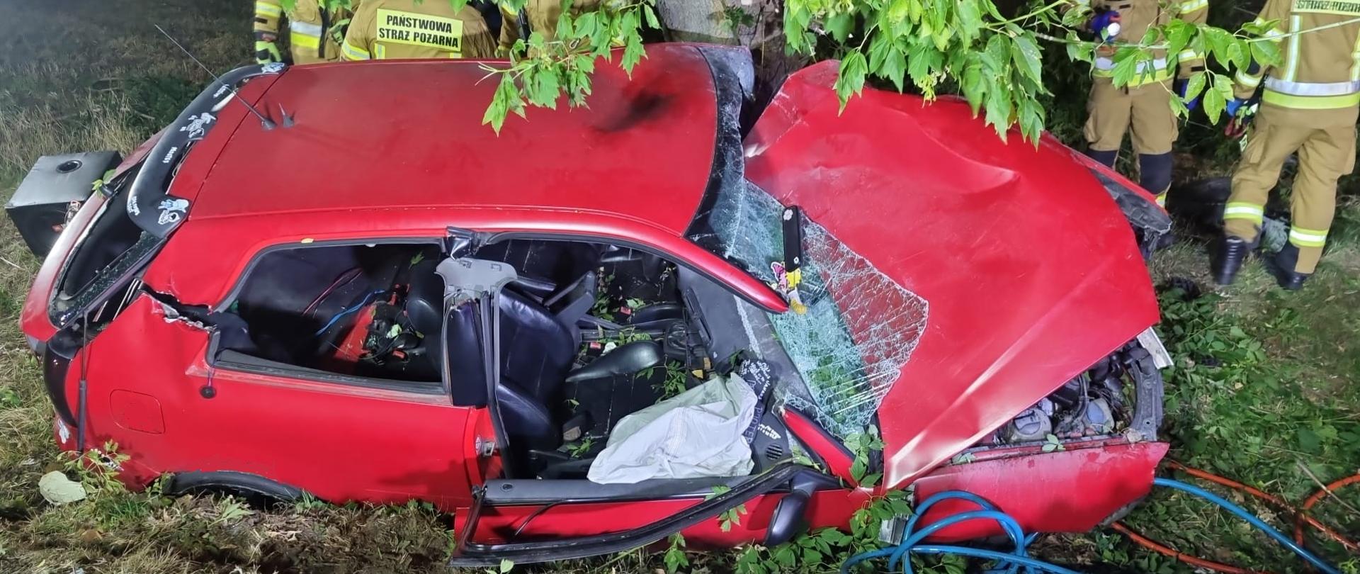 Czerwony samochód osobowy na poboczu drogi DK11. Dach usunięty przez strażaków, Widoczne węże od narzędzi hydraulicznych. Na ziemi hełm strażacki i koc. Na drugim planie strażacy. 