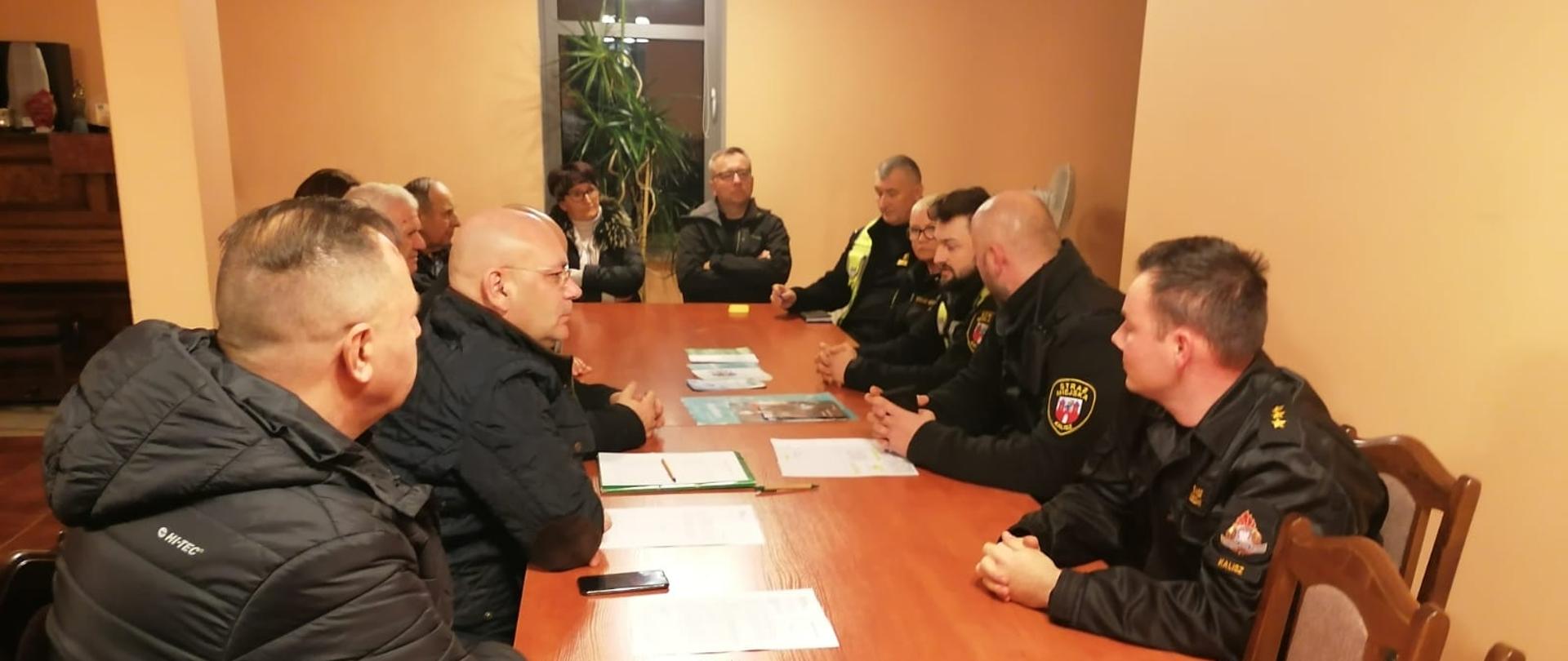 Zdjęcie wykonane wewnątrz pomieszczenia. Światło sztuczne. Przy stole osoby, przedstawiciele służb mundurowych. Stół drewniany, brązowy.