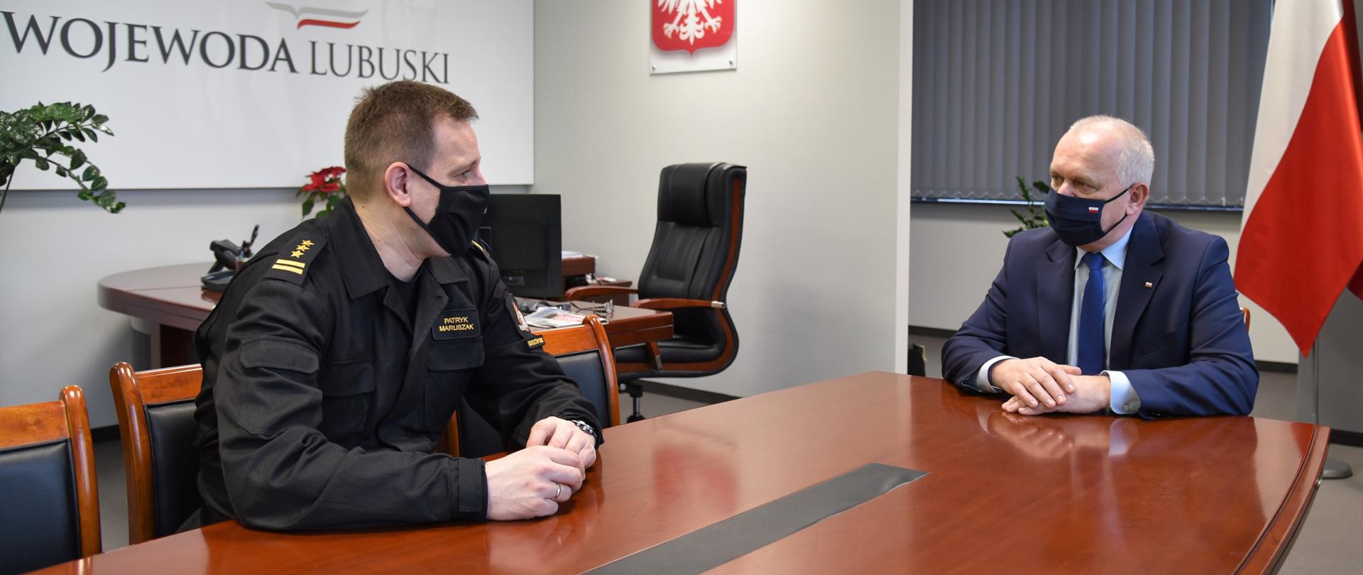 Wojewoda siedzi u szczytu stołu, a komendant w mundurze po jego prawej stronie. W tle widać burko z odsuniętym krzesłem, biało-czerwoną flagę i kwiaty 
