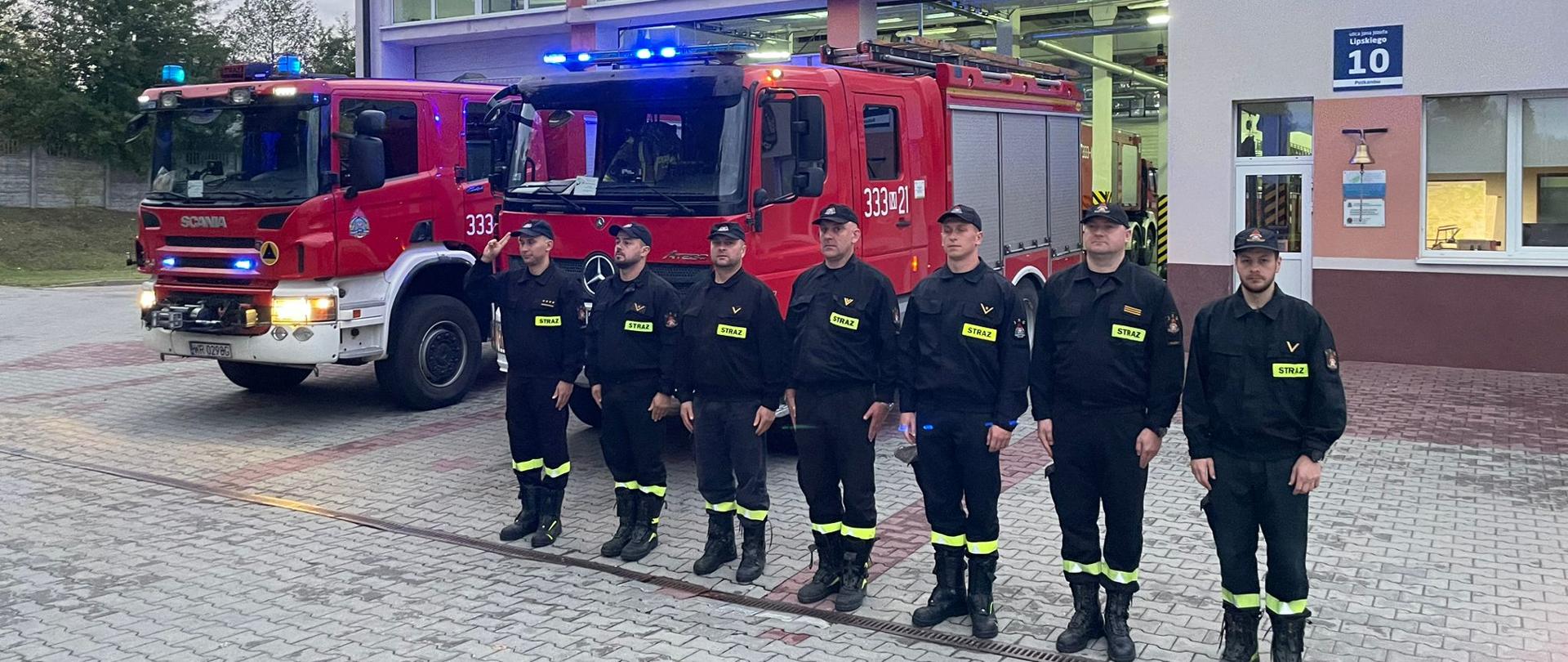 Zdjęcie wykonane przed jednostką ratowniczo – gaśniczą numer 3 w Radomiu. Na zdjęciu widać strażaków ustawionych w szeregu przed jednostką. Strażacy ubrani w ubrania koszarowe czarnym z jaskrawymi elementami odblaskowymi. Za nimi ustawione są pojazdy ratowniczo – gaśnicze. Pojazdy mają włączone alarmowe sygnały świetlne i dźwiękowe. Strażacy oddają hołd zmarłemu koledze strażakowi.