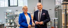 Wiceminister Mrówczyńska w niebieskiej marynarce stoi obok mężczyzny w czarnym garniturze, za nimi widać metalowe regały na których stoją różne naczynia.