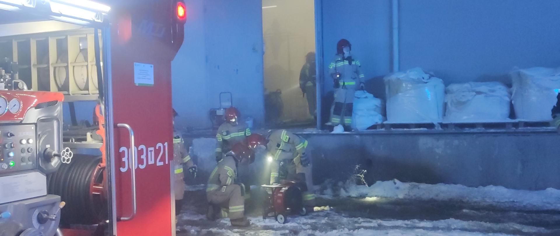 Zdjęcie przedstawia halę produkcyjną. Przez otwarte drzwi widać silne zadymienie wewnątrz. Poniżej rampy rozładunkowej strażacy przygotowują wentylator osiowy do oddymiania pomieszczenia.