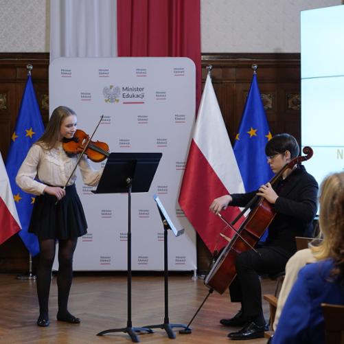 Zdjęcie przedstawia duet skrzypcowo wiolonczelowy podczas wykonania utworu, w tle ekran multimedialny, flagi Polski i unii europejskiej oraz baner minister edukacji