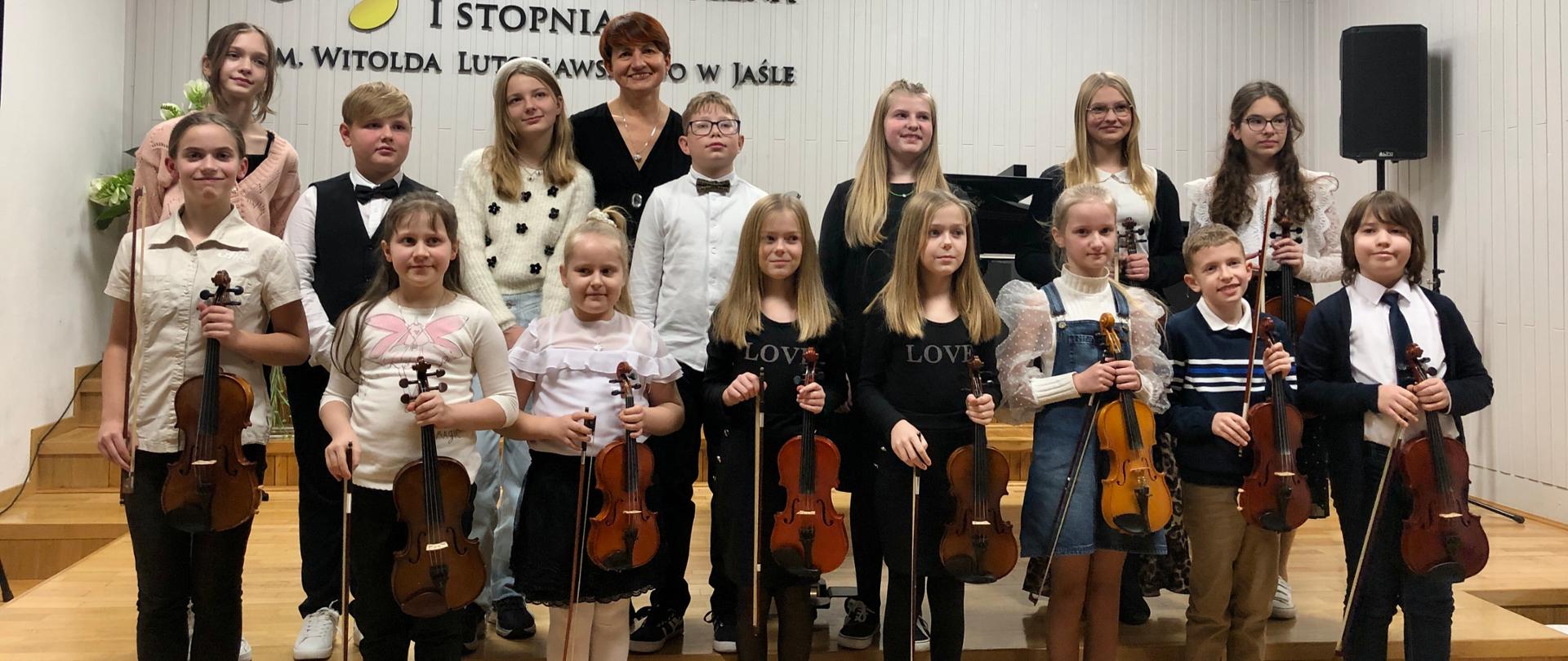 Grupa uczniów oraz pedagog w strojach galowych. Uśmiechnięci trzymają w rękach swoje instrumenty. W tle Godło oraz logo szkoły. Ogólna kolorystyka – biało-czarna.