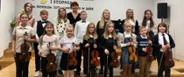 Grupa uczniów oraz pedagog w strojach galowych. Uśmiechnięci trzymają w rękach swoje instrumenty. W tle Godło oraz logo szkoły. Ogólna kolorystyka – biało-czarna.
