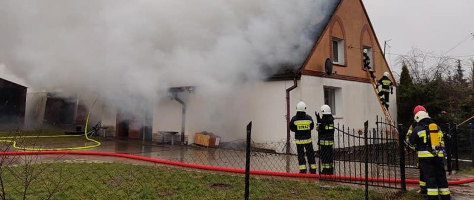 Widok na budynek objęty pożarem. Budynek niski, dach dwuspadowy. Z budynku unoszą się kłęby dymu. Połowa budynku zakryta gęstym dymem. Dym ścieli się bardzo nisko. Przy budynku strażacy prowadzący działania.