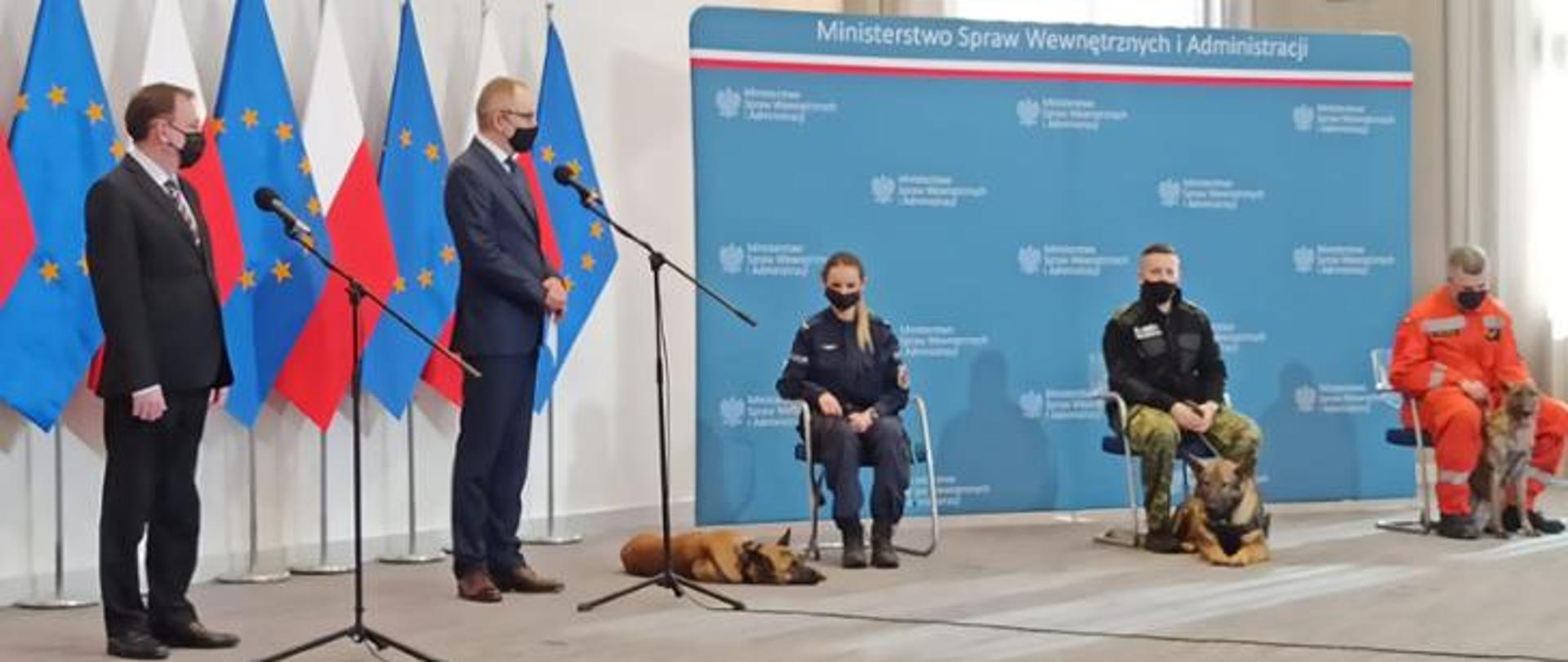 Konferencja przedstawicieli rządu w sprawie opieki nad zwierzętami. Dwóch mężczyzn stoi przy mikrofonach odpowiadając na pytania dziennikarzy po prawej stronie przedstawiciele służb na krzesłach (kobieta i dwóch mężczyzna) przy nich znajdują się psy szkolone do pomocy w tle flagi. 