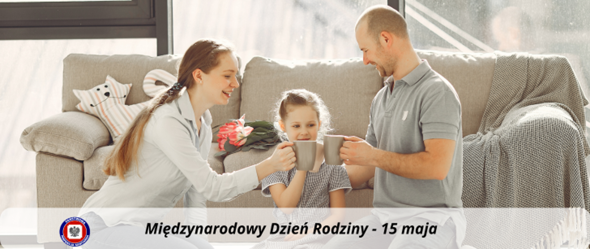 Na podłodze w salonie na tle szarej kanapy klęczą kobieta, mężczyzna i dziecko, stukają się kubkami. Poniżej, na jasnym półprzezroczystym pasku ciemny napis Międzynarodowy Dzień Rodziny - 15 maja. Z jego lewej strony logo Państwowej Inspekcji Sanitarnej.