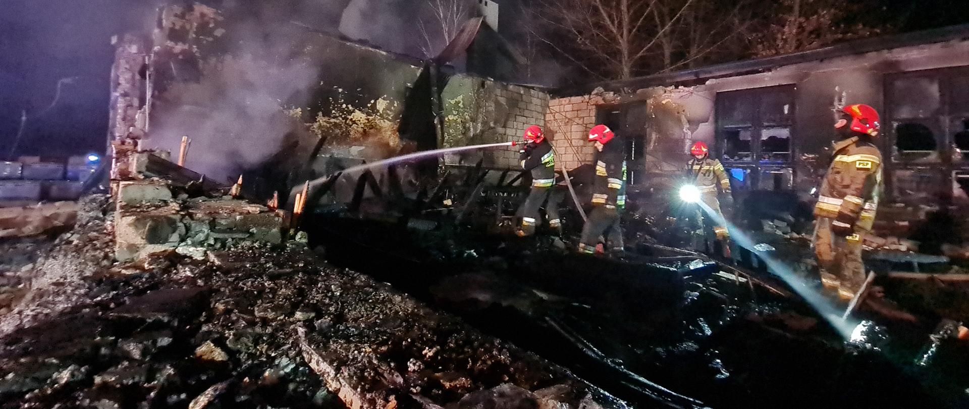 Zdjęcie przestawia funkcjonariuszy Państwowej Straży Pożarnej oraz Ochotniczej Straży Pożarnej podczas działań gaśniczych. W tle pogorzelisko remizy - zniszczony obiekt. 