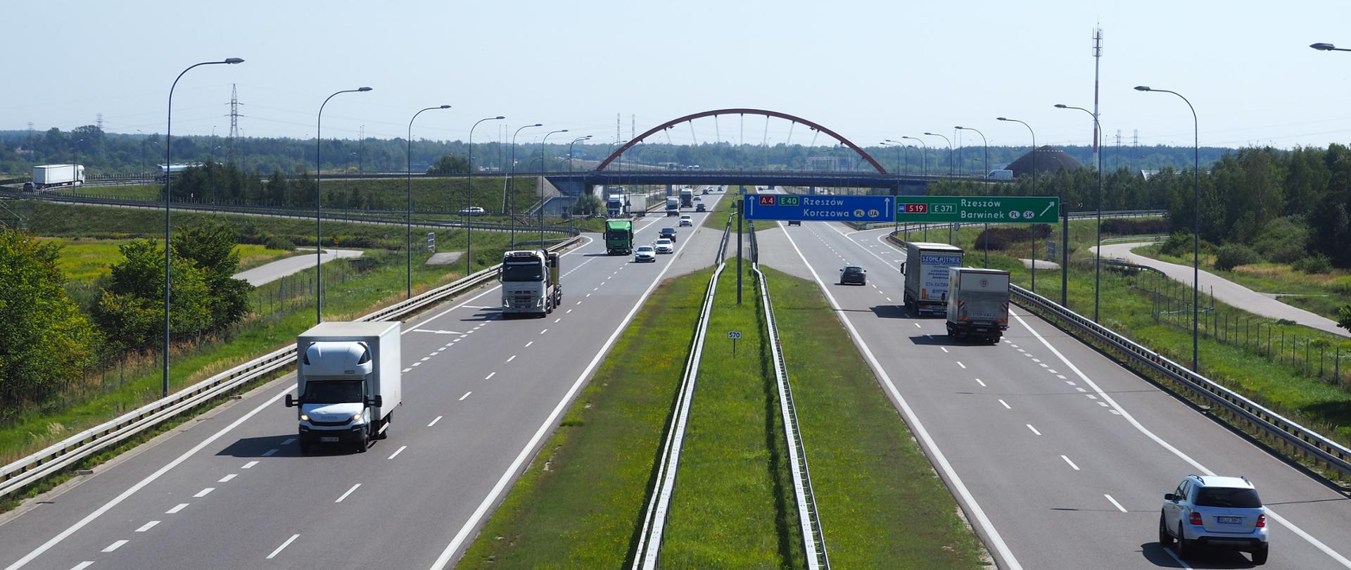 Dwujezdniowa droga szybkiego ruchu z umiarkowanym ruchem samochodów i ciężarówek, widok z góry; w tle wiadukt i znaki drogowe.