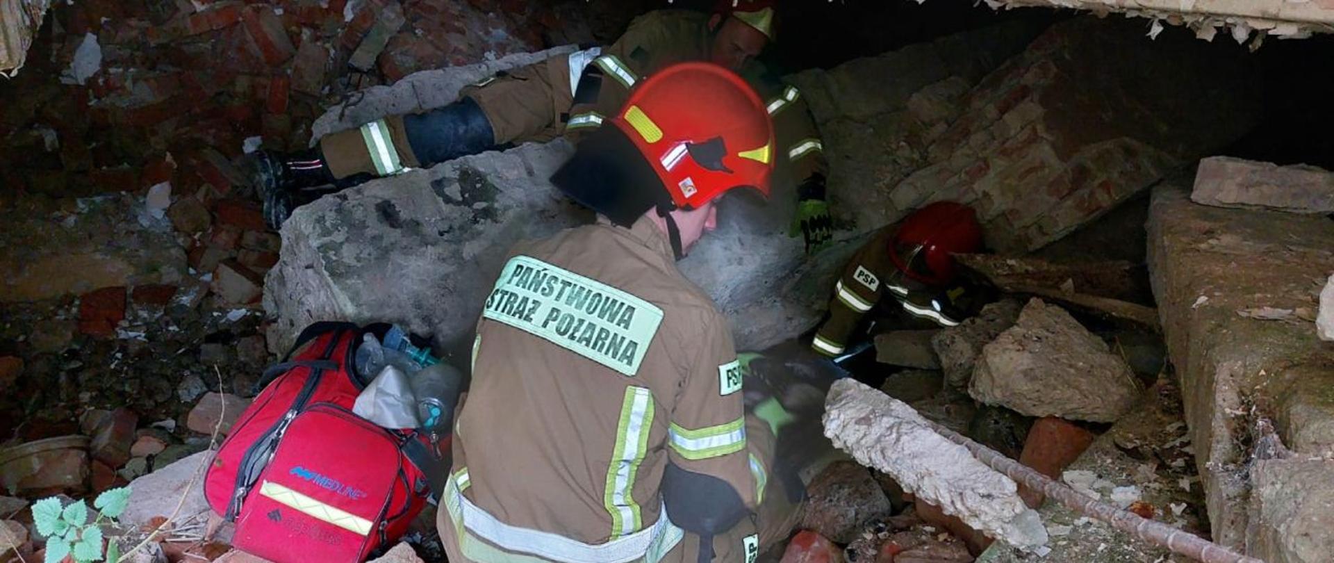 Strażacy Państwowej Straży Pożarnej w hełmach koloru czerwonego i ubraniach specjalnych działają na gruzowisku betonowo-ceglanym realizują kwalifikowaną pierwszą pomoc u osoby poszkodowanej znajdującej się w gruzowisku. Działanie przeprowadzone w ramach szkolenia z działań poszukiwawczo-ratowniczych.