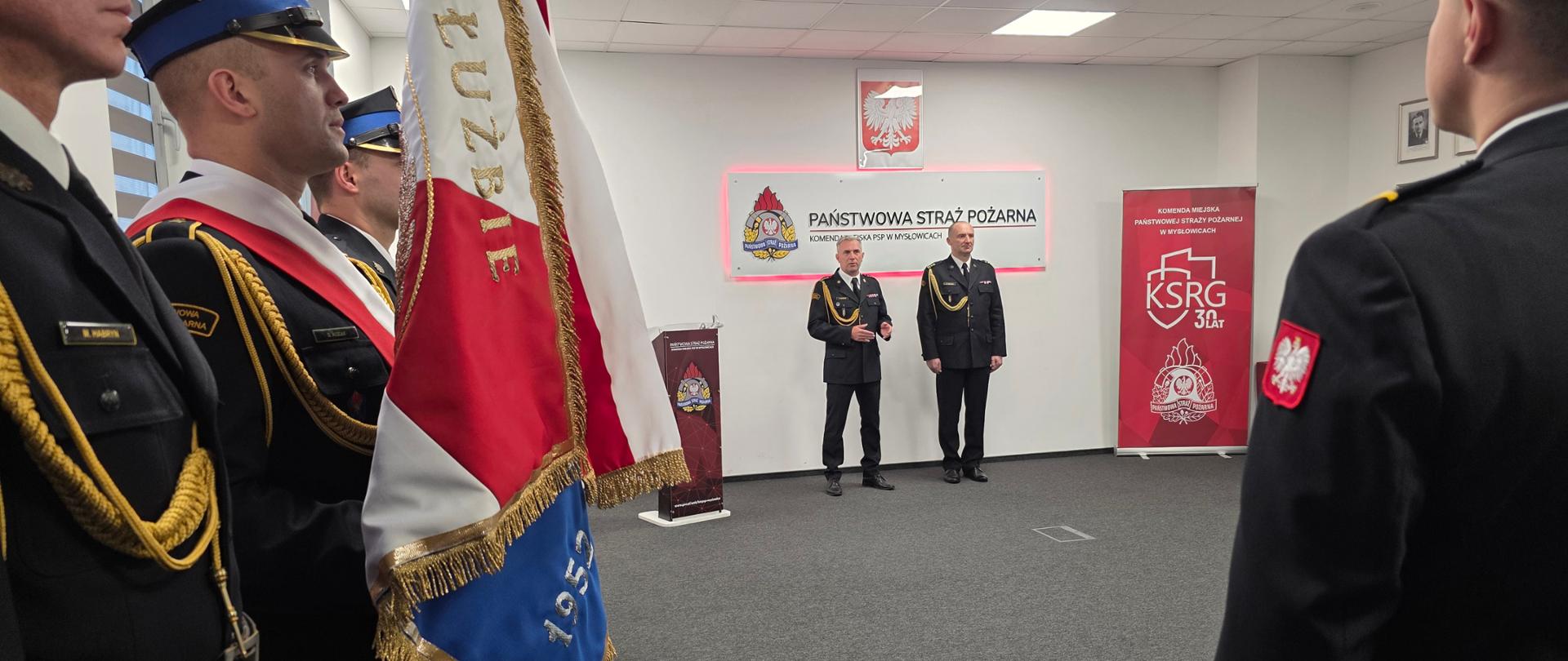 Zdjęcie przedstawia scenę z uroczystej zbiórki w Sali Sztabowej Komendy Miejskiej Państwowej Straży Pożarnej w Mysłowicach, koncentrując się na poczcie sztandarowym i dowództwie.
Poczet Sztandarowy (Pierwszy Plan): Na pierwszym planie, po lewej stronie kadru, widoczny jest fragment sztandaru, z widocznymi barwami biało-czerwonymi i haftowanymi napisami oraz cyframi. Obok sztandaru stoją strażacy w galowych mundurach, w tym jeden w szarfie (prawdopodobnie dowódca pocztu) i kołnierzu. Ich obecność nadaje scenie bardzo podniosły i oficjalny charakter.
Dowództwo (Tło): W centrum zdjęcia, w tle, stoją dwaj mężczyźni w ciemnych, galowych mundurach PSP, prawdopodobnie Komendant Miejski PSP w Mysłowicach i jego zastępca lub zaproszony gość, którzy przemawiają lub obserwują przebieg uroczystości. Jeden z nich gestykuluje w trakcie przemówienia.
Tło i Dekoracje: W tle, na ścianie, widać te same elementy dekoracyjne co na poprzednim zdjęciu: duży, biało-czerwony panel "PAŃSTWOWA STRAŻ POŻARNA KOMENDA MIEJSKA PSP W MYSŁOWICACH", a po prawej banner okolicznościowy "KSRG 30 LAT". Nad centralnym panelem wisi godło państwowe.
Zdjęcie podkreśla rangę uroczystości związanej z Narodowym Świętem Niepodległości i wręczaniem awansów/wyróżnień, a obecność sztandaru akcentuje tradycję i ciągłość służby.