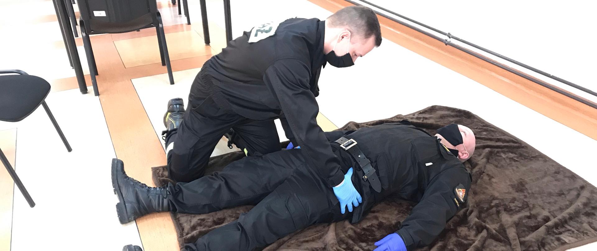 Na zdjęciu trzecim strażak-ratownik wykonuje symulację badania urazowego osoby poszkodowanej. Badanie rozpoczyna od głowy, następnie bada szyję, obojczyki, klatkę piersiową, brzuch, miednicę, kończyny dolne, kończyny górne oraz plecy i pośladki. Badanie urazowe polega na szybkim zidentyfikowaniu krwawień oraz innych nieprawidłowości poszczególnych części ciała.