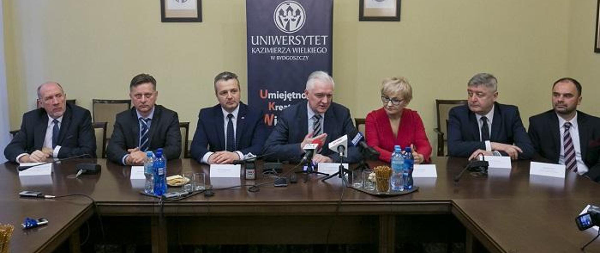 Minister Gowin i przedstawiciele władz uczelni siedzą na tle ściany z dyplomami i plakatem UKW