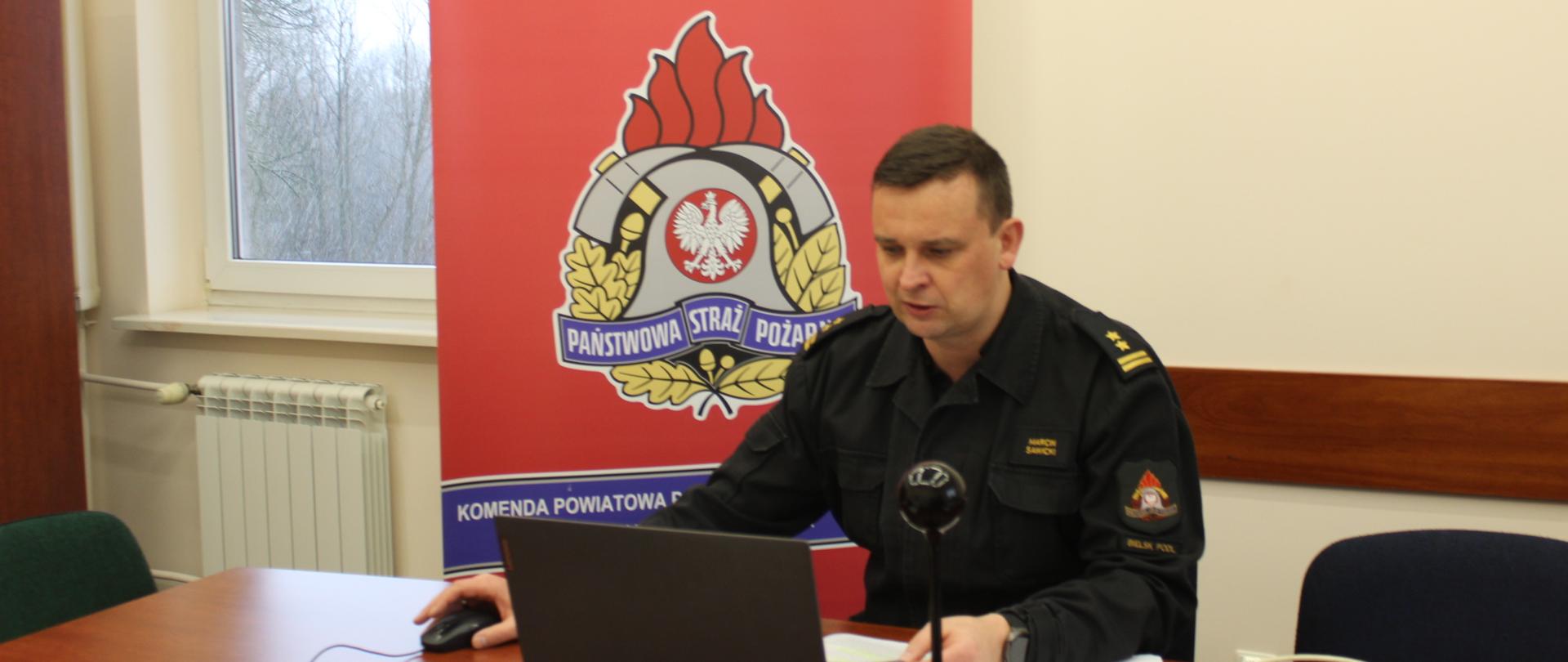Zdjęcie przedstawia Funkcjonariusza Państwowej Straży Pożarnej w czarnym mundurze przed komputerem z tyłu logo Państwowej Straży Pożarnej na czerwonym tle