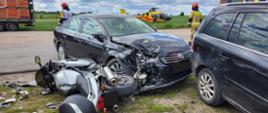 Wypadek drogowy w Koziołkach, zderzenie motocykla z samochodem osobowym, motocykl leży na poboczu obok samochodu, który ma uszkodzony przód w wyniku powstałego zdarzenia, w pobliżu pojazdów leżę szczątki uszkodzonych elementów, przed uszkodzonym pojazdem widać zaparkowany inny samochód, w oddali na polu widać śmigłowiec lotniczego pogotowia ratunkowego, na poboczu za drogą stoi samochód ciężarowy, dwóch strażaków w ubraniach specjalnych i hełmach prowadzą działania ratownicze.