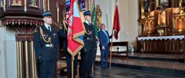 Zdjęcie przedstawia grupę mężczyzn stojących wewnątrz bogato zdobionego kościoła. Na pierwszym planie po lewej stronie widoczna jest asysta honorowa składająca się z trzech strażaków w czarnych garniturach galowych, rogatywkach oraz rękawiczkach. Jeden z nich trzyma sztandar. Za nimi, nieco głębiej w nawie, stoi mężczyzna w niebieskim garniturze, który również podtrzymuje czerwony sztandar. Cała grupa stoi w postawie zasadniczej, zwracając się w stronę ołtarza.
Tło kadru ukazuje tradycyjne wnętrze sakralne z elementami architektury gotyckiej lub neogotyckiej. Po lewej stronie znajduje się fragment rzeźbionej, ciemnej ambony wspartej na ozdobnych kolumnach. Po prawej stronie widoczny jest ołtarz główny z figurami świętych oraz tabernakulum, przed którym stoi stół ołtarzowy nakryty białym obrusem z koronką. Światło do wnętrza wpada przez witraże umieszczone wysoko w ścianie bocznej kościoła, oświetlając kamienną posadzkę.