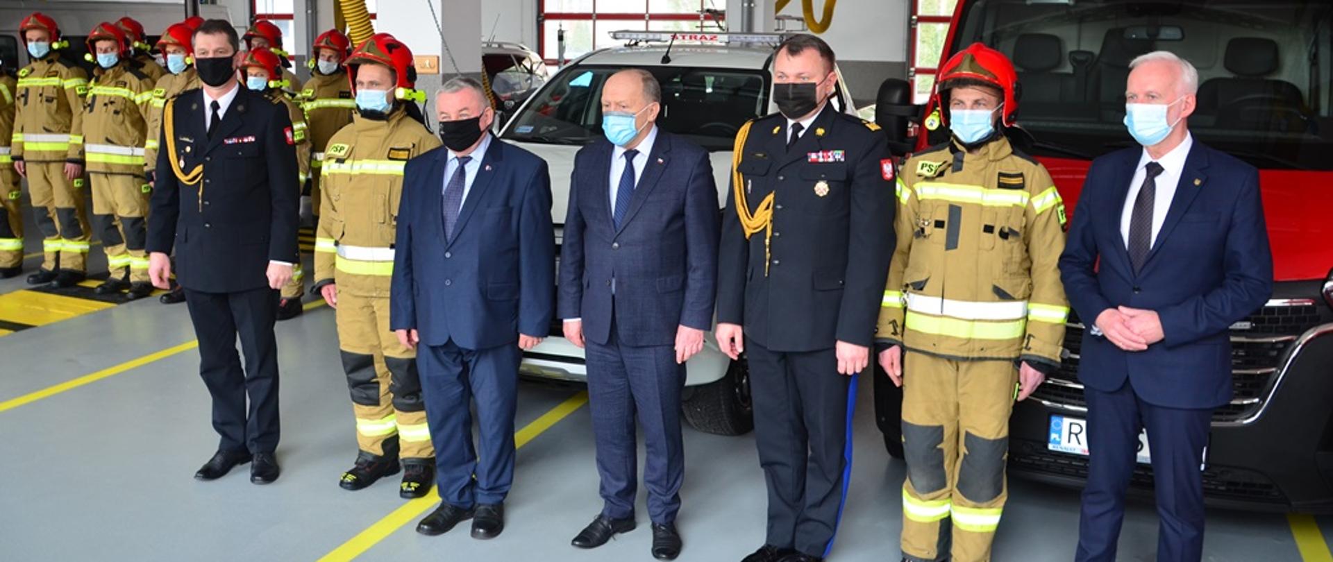 Mężczyźni stoją w postawie zasadniczej w szeregu na garażu przed dwoma samochodami pożarniczymi. Dwóch z nich występuje w umundurowaniu wyjściowym, dwóch w umundurowaniu bojowym, a pozostali w ubraniach cywilnych. W głębi widoczni strażacy w umundurowaniu bojowym, którzy stoją w dwuszeregu. 