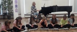 W sali koncertowej na podłodze siedzi grupa dziewcząt i gra na leżących przed nimi cymbałkach. Za nimi przy fortepianie siedzi kobieta i akompaniuje im na fortepianie. 