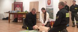 Kobieta ćwiczy zabiegi resuscytacyjne na fantomie. Uciska klatkę piersiową. Strażacy prowadzą instruktarz