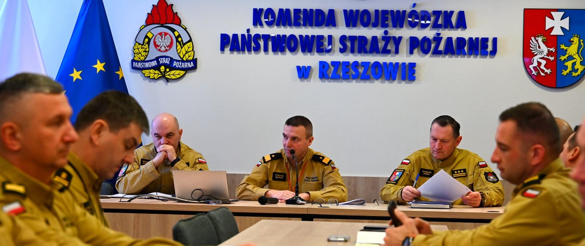 Widok na salę konferencyjną Komendy Wojewódzkiej PSP w Rzeszowie. Na pierwszym planie siedzi oficer Państwowej Straży Pożarnej w piaskowym umundurowaniu służbowym, trzymający w prawej dłoni mikrofon i przemawiający do zgromadzonych. Przed nim na stole leżą dokumenty i notes. Obok siedzi drugi oficer, pochylony nad notatkami. W tle widoczne są elementy wystroju sali oraz godła i symbole instytucji.