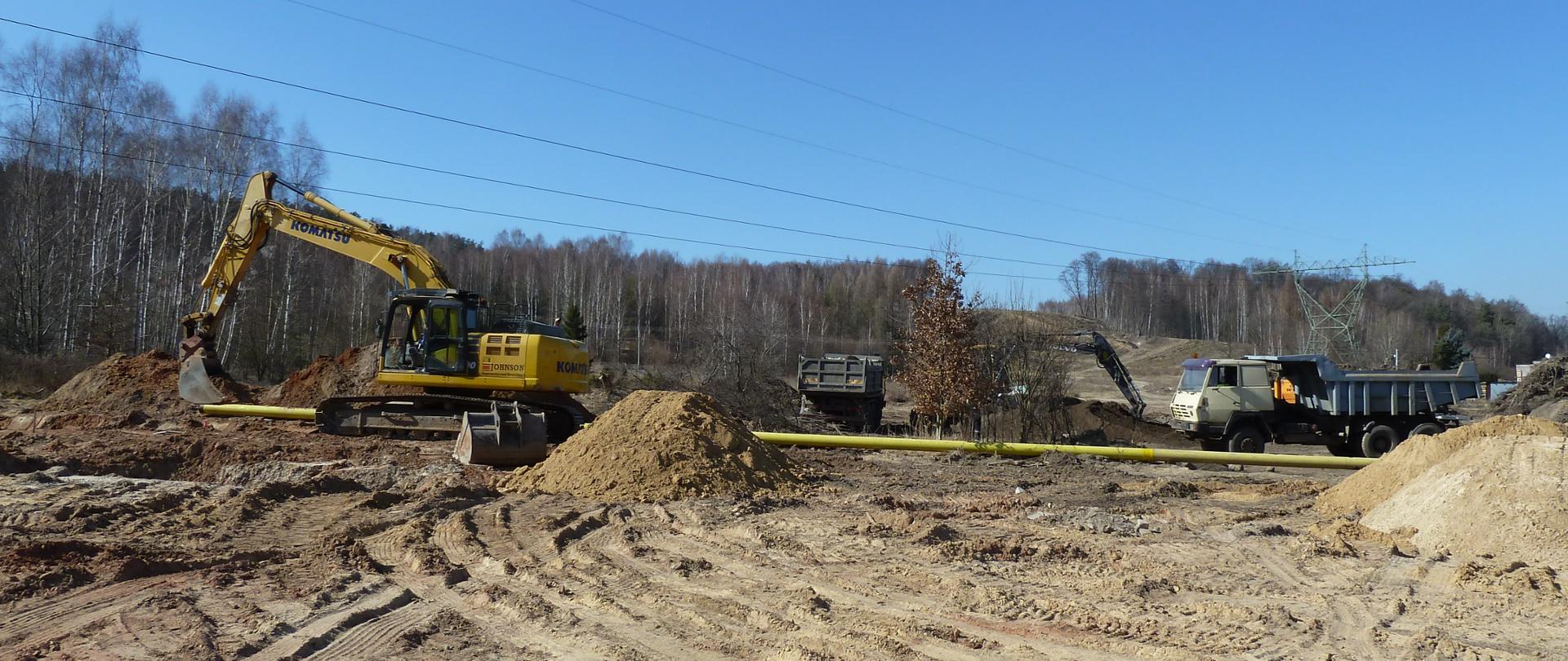 DK42 obwodnica Wąchocka - roboty ziemne w śladzie przyszłej drogi między drzewami, koparka, dwa samochody ciężarowe wywrotki, żółta rura w poprzek drogi 