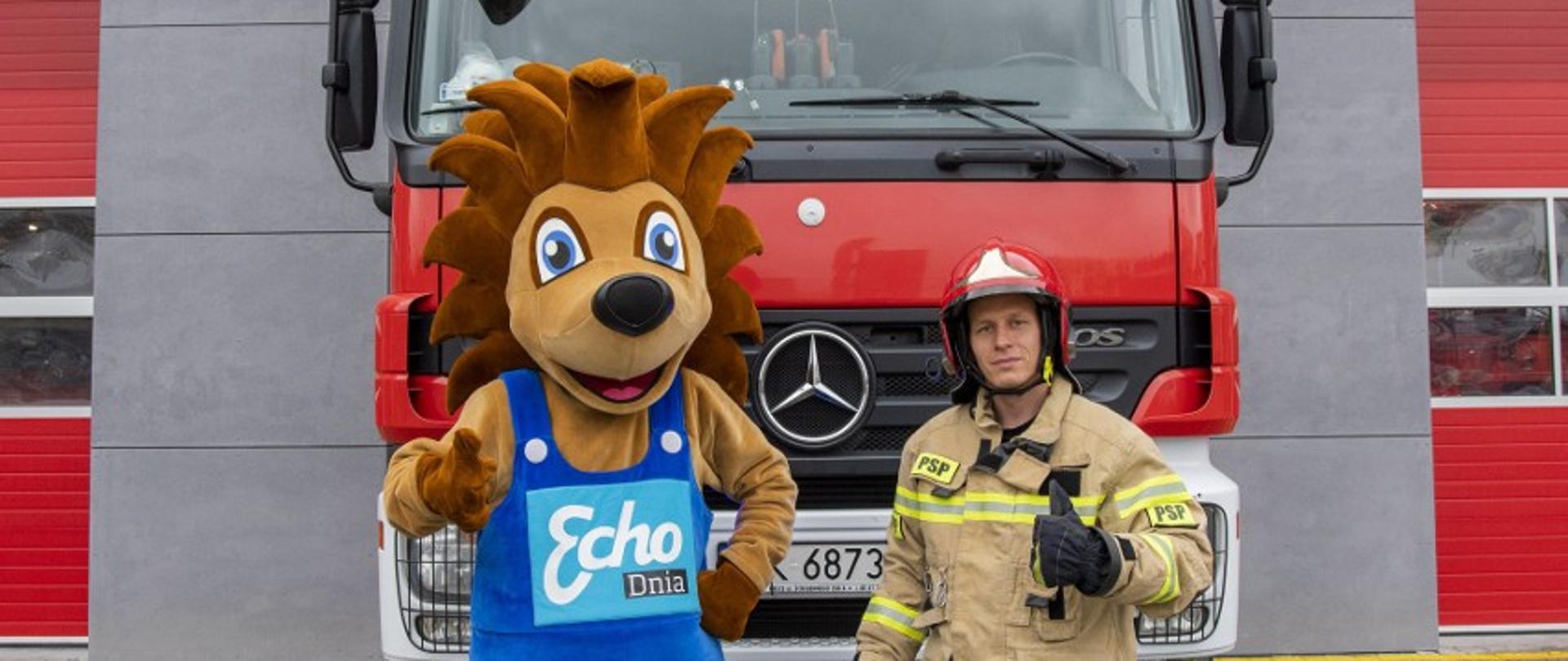 Na zdjęciu znajduje się strażak razem jeżem z akcji Echa dnia. Stoją przed samochodem pożarniczym. W tle widać strażnicę JRG 1.