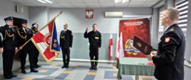 Strażak w trackie składnia ślubowania. Dwa palce podniesione ma ku górze. Komendant odczuje kolejne wersy treści ślubowania. Z prawej stroni widoczny sztandar komendy. 
