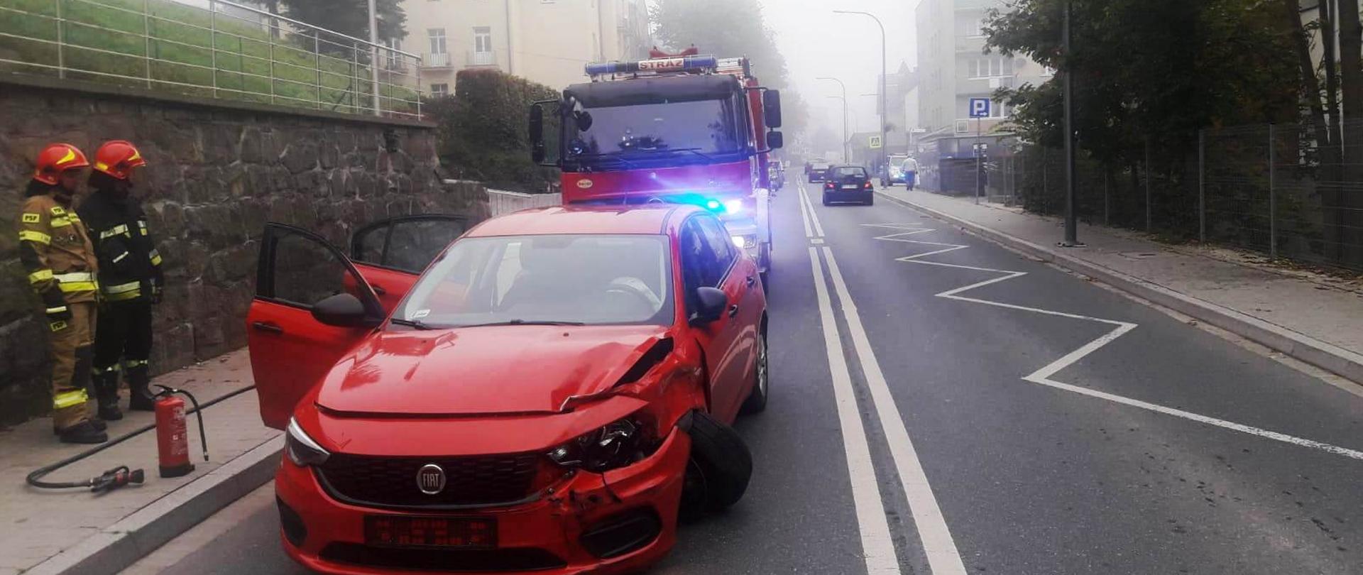 Uszkodzony samochód na jednym pasie. Obok niego strażacy a za nim samochód pożarniczy