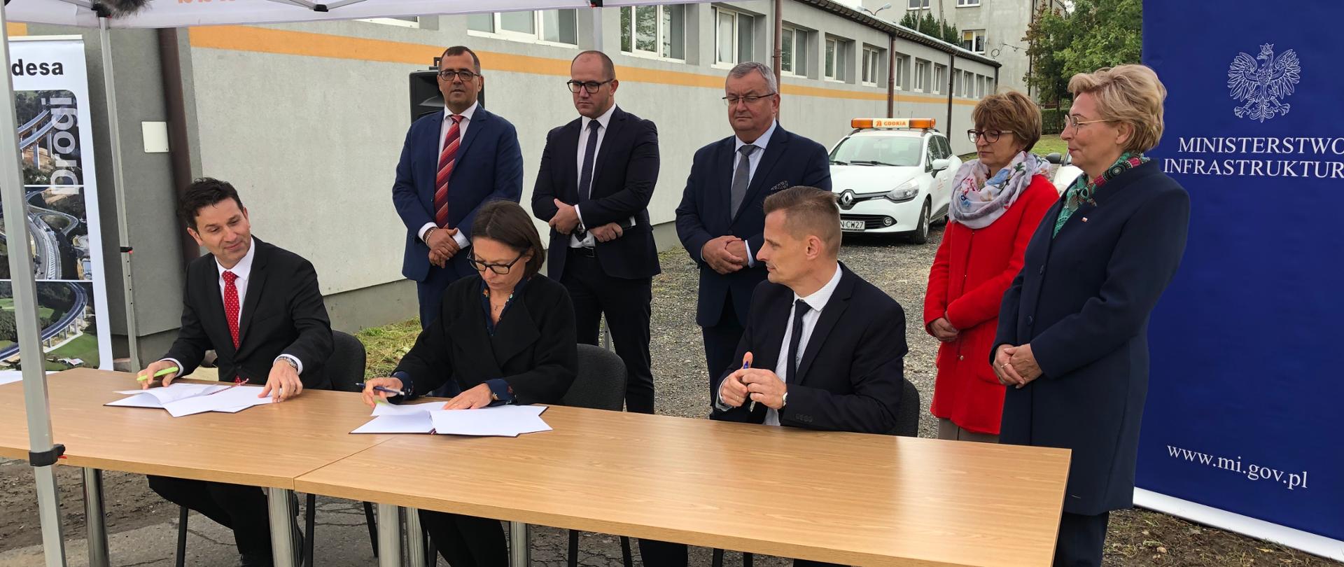 Fotografia przedstawia uroczystość podpisania umowy na zaprojektowanie i budowę ostatniego z trzech odcinków drogi ekspresowej S7 między Płońskiem a Czosnowem. Na pierwszym planie 3 osoby - kobieta i dwóch mężczyzn - podpisują umowę, siedząc za stołem. Tuż za nimi stoi 5 osób - 3 mężczyzn i 2 kobiety. W tle ścianka wizerunkowa Ministerstwa Infrastruktury, budynek oraz pojazd techniczny Generalnej Dyrekcji Dróg Krajowych i Autostrad
