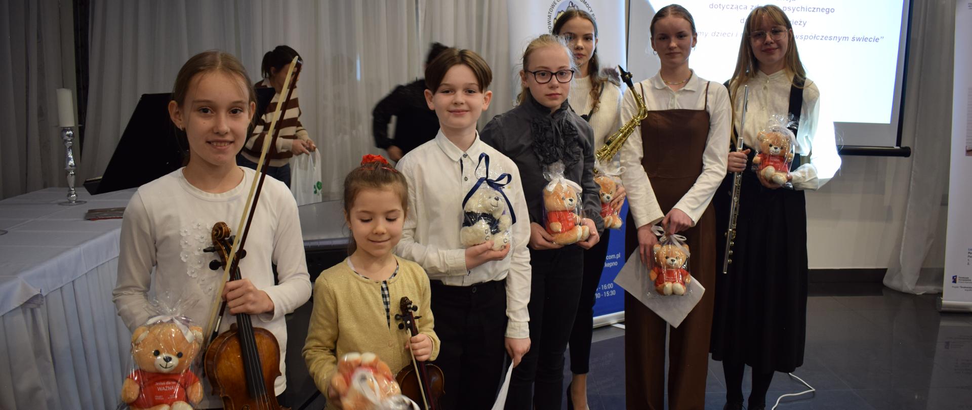 Zdjęcie przedstawia grupę uczniów ubranych na galowo i trzymających w dłoniach pluszowe maskotki. Pierwsze dwie uczennice od lewej trzymają ponadto w dłoniach skrzypce, a dalej dziewczynka trzyma saksofon i ostatnia w szeregu flet. W tle od lewej widać stół przykryty białym obrusem, na którym stoi świecznik, dalej pulpit do nut, okna zasłonięte firanami i rozwinięty na statywie ekran z wyświetlonym logo powiatu kępińskiego i powiatowego centrum pomocy rodzinie oraz tematem konferencji. 