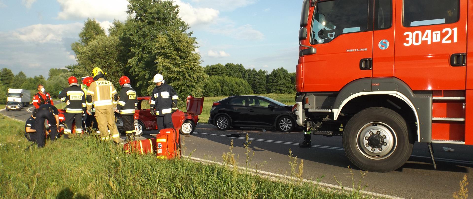 Zdjęcie przedstawia strażaków udzielającym pomocy poszkodowanym w zdarzeniu. Za nimi znajdują się rozbite pojazdy. Z prawej strony widać samochód straży pożarnej.