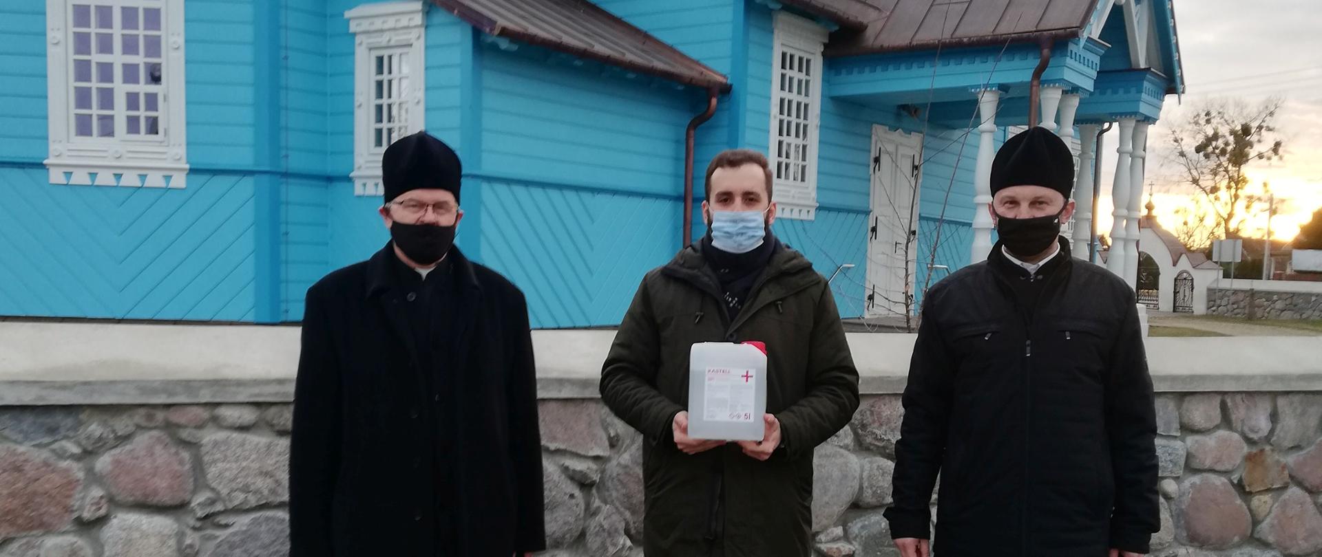Na tle niebieskiej cerkwi stoi trzech duchownych w sutannach. Jeden z nich stojący po środku trzyma zbiornik z płynem do dezynfekcji.