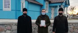 Na tle niebieskiej cerkwi stoi trzech duchownych w sutannach. Jeden z nich stojący po środku trzyma zbiornik z płynem do dezynfekcji.
