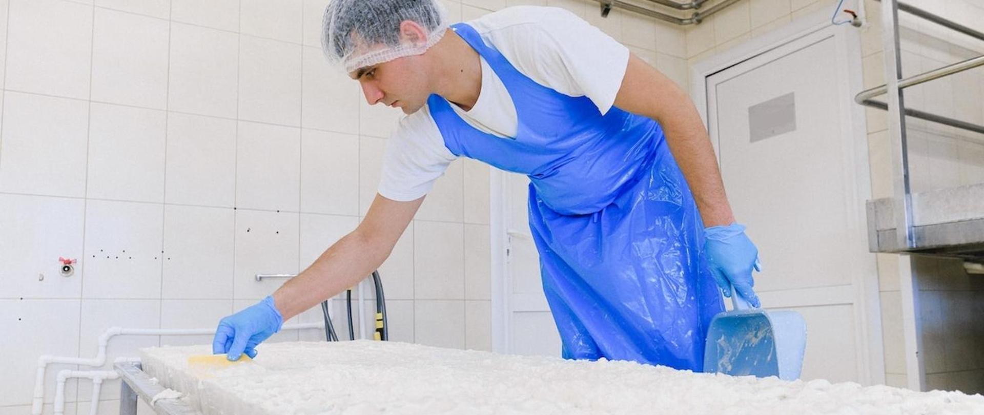 Mężczyzna w białej koszulce z nałożonym niebieskim fartuchem pracuje PRZY TAŚMIE PRODUKCYJNEJ