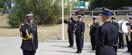 Komendant główny Państwowej Straży Pożarnej oddaje honor stojąc przed strażakiem ochotniczej straży pożarnej. Niedaleko stoi orkiestra Morskiego Oddziału Straży Granicznej która gra. 