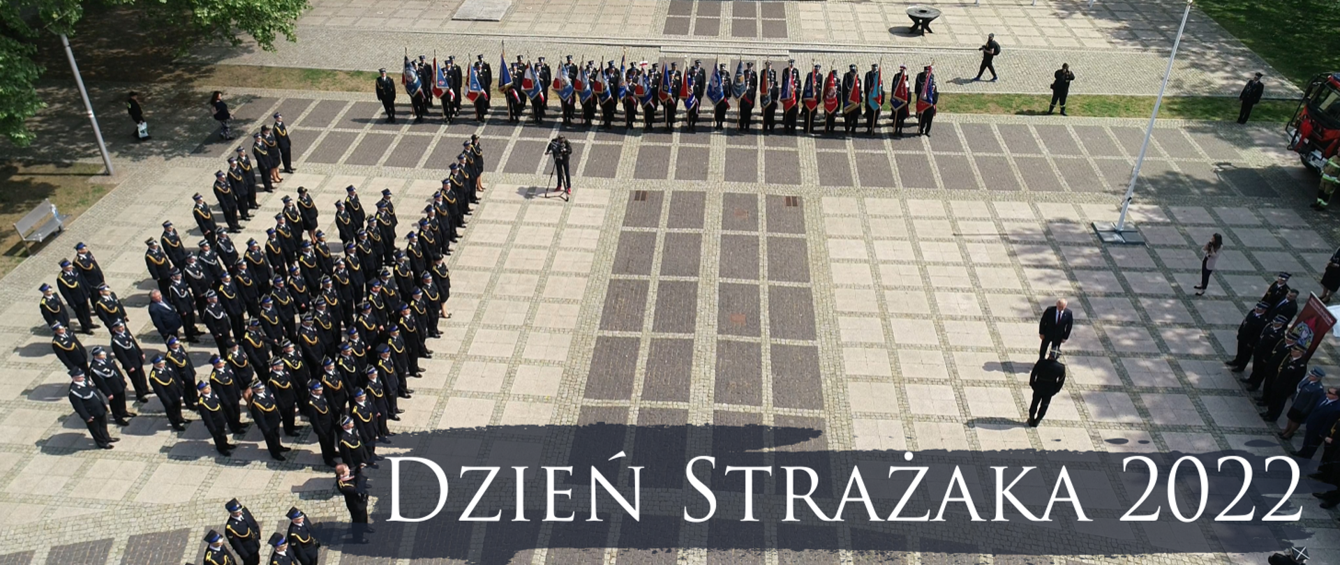 Fotografia przedstawia ujęcie z góry na plac na którym ustawione są pododdziały strażaków w mundurach galowych. Po lewej stronie kadru stoją odznaczeni i awansowani, w górnej części widoczny szereg pocztów sztandarowych, po prawej stronie uchwycony moment składania meldunku przez dowódcę uroczystości.