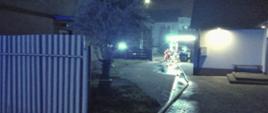 Zdjęcie przedstawia posesję, gdzie doszło do pożaru stodoły. W głębi widać ratowników do których dochodzi wąż pożarniczy.