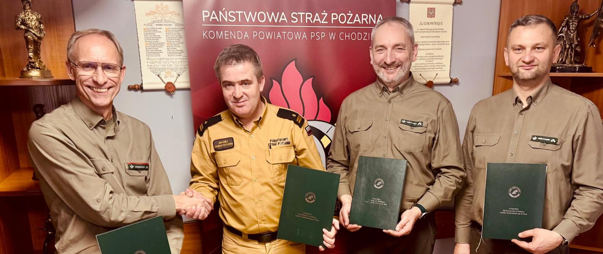 Zdjęcie przedstawia leśników i strażaka podczas podpisywania porozumienia w sprawie utworzenia Zintegrowanego Punktu Alarmowo-Dyspozycyjnego.
W tle samochody pomieszczenie.
