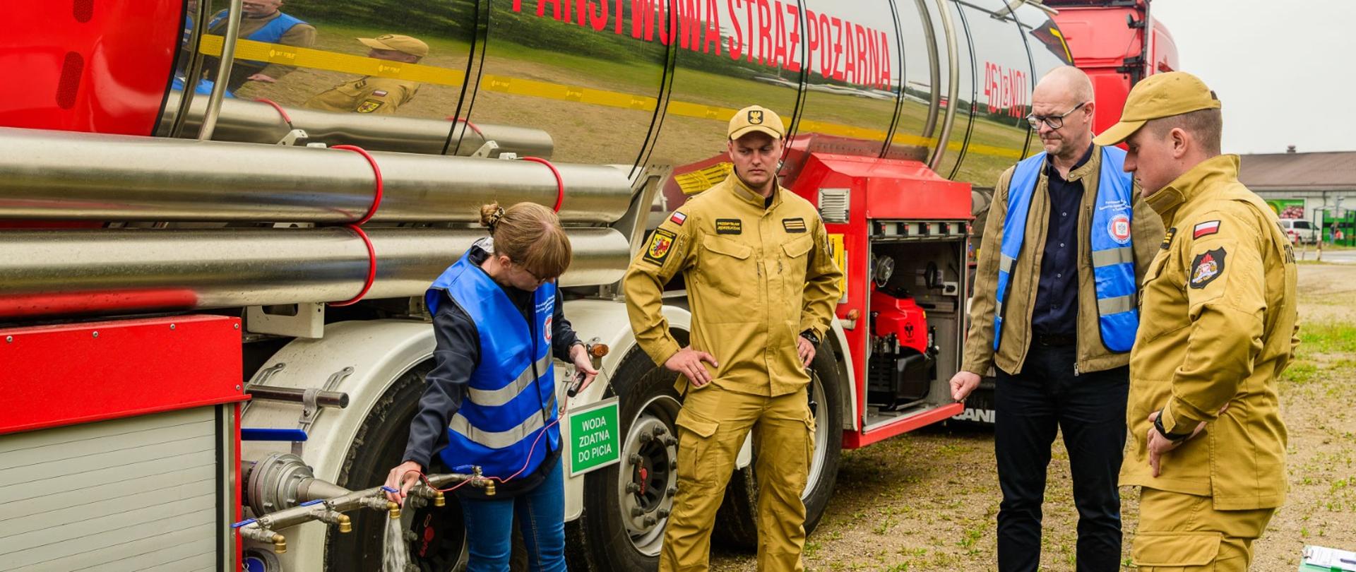 Kobieta w niebieskiej kamizelce otwiera zawór przy dużej cysternie z napisem „Państwowa Straż Pożarna”, z którego na ziemię leje się woda. Sytuację obserwuje dwóch strażaków w beżowych mundurach oraz mężczyzna w okularach i niebieskiej kamizelce.