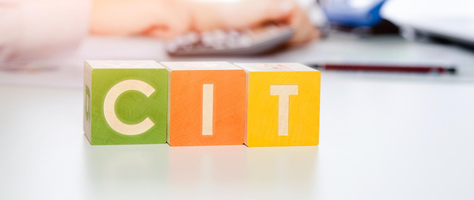 Trzy różnokolorowe klocki, na nich napis CIT, w tle przybory biurowe