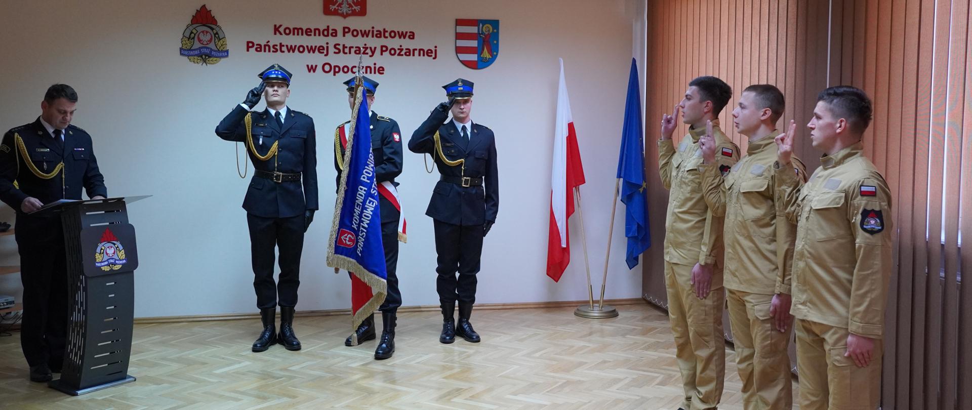 Sala Komendy Powiatowej PSP, zdjęcie przedstawia moment ślubowania na sztandar strażacy z uniesionymi ramionami, naprzeciwko Komendant Powiatowy stojący za mównicą i funkcjonariusze komendy. W tle flagi koloru biało-czerwonego i niebieskiego oraz godło Polski, logo Państwowej Straży Pożarnej i logo Powiatu Opoczyńskiego.